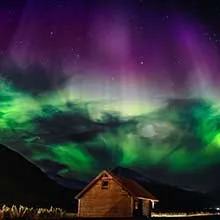 Green and purple northern lights shine over a starry night sky above a small wooden cabin nestled between mountains.