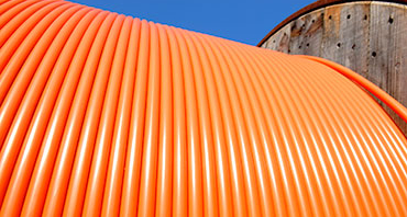 Close-up, low-angle view of a large spool wrapped with bright orange, ribbed cable, set against a blue sky and a weathered industrial tank.