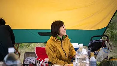 A woman with short dark hair, wearing a yellow jacket, smiles while sitting at a campsite beneath a large yellow and green tarp.