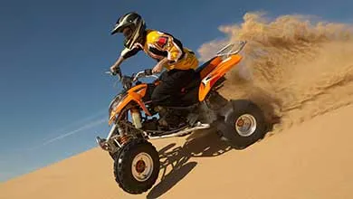 A person wearing a helmet rides an orange ATV up a steep sand dune, kicking up sand under a clear blue sky.
