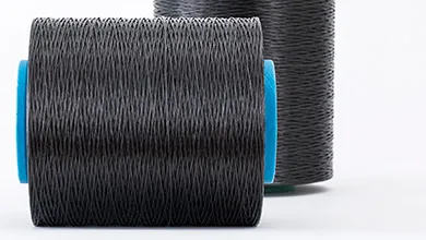 Close-up of a dark gray, textured thread spool lying on its side with a blue end visible; another spool stands upright in the background.