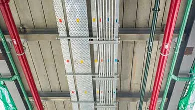 Low-angle view of an industrial ceiling with red and green pipes next to silver electrical conduits and perforated cable trays.