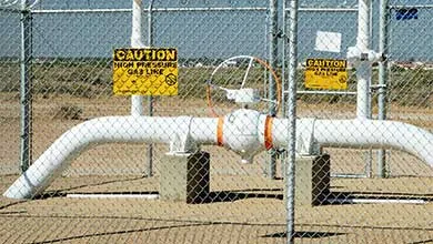 White industrial pipes with a central valve are secured behind a chain-link fence displaying yellow "Caution High Pressure Gas Line" signs. The setup is in a dry, open area with hills visible in the distance under a clear sky.