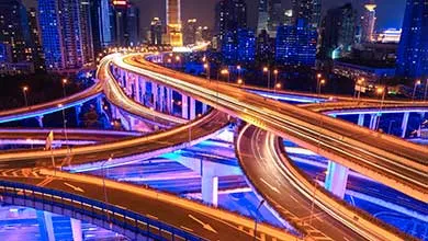 A multi-level highway interchange at night, with orange and white light trails from cars. The city skyline and overpasses are lit in blue and purple.
