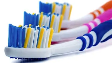 Close-up of a toothbrush with blue, white, and yellow bristles, with several colorful toothbrushes lined up behind it on a white background.