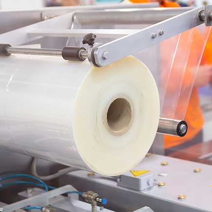 Close-up of a large roll of clear plastic film being fed into an industrial packaging machine.